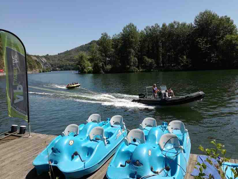 A serene river scene features bright blue pedal boats lined along a wooden dock. A small motorboat is seen cruising on the water while tree-covered hills rise in the background, creating a calming natural landscape.