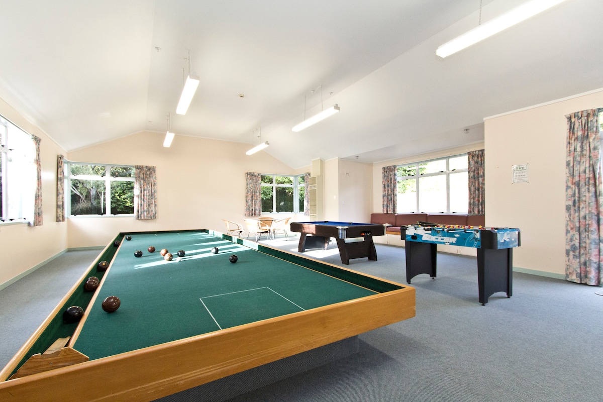 A spacious recreation room features a pool table and a large indoor bowling area. Natural light enters through multiple windows, creating a bright atmosphere. Comfortable seating is arranged in the distance, complemented by curtains with floral patterns.
