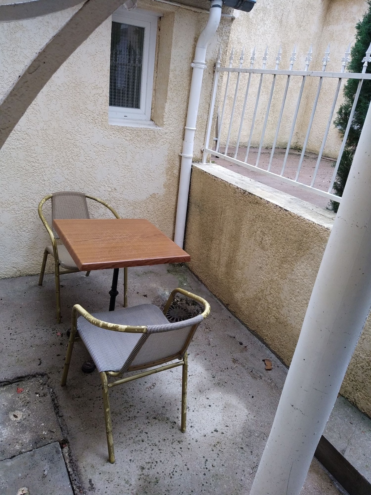 A small outdoor seating area is featured, consisting of a wooden table with two metal-framed chairs. The space is enclosed by a low wall, providing privacy. Natural light filters through, creating a cozy setting in a peaceful corner.