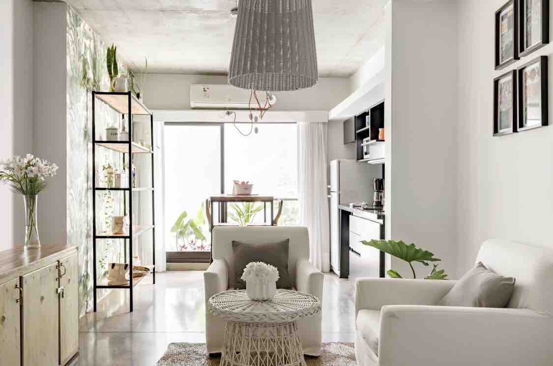 A modern living space features two comfortable armchairs positioned around a circular coffee table. A light fixture hangs above, and a shelving unit displays plants and decor. Large glass doors allow natural light to fill the room, leading to a small balcony with greenery.