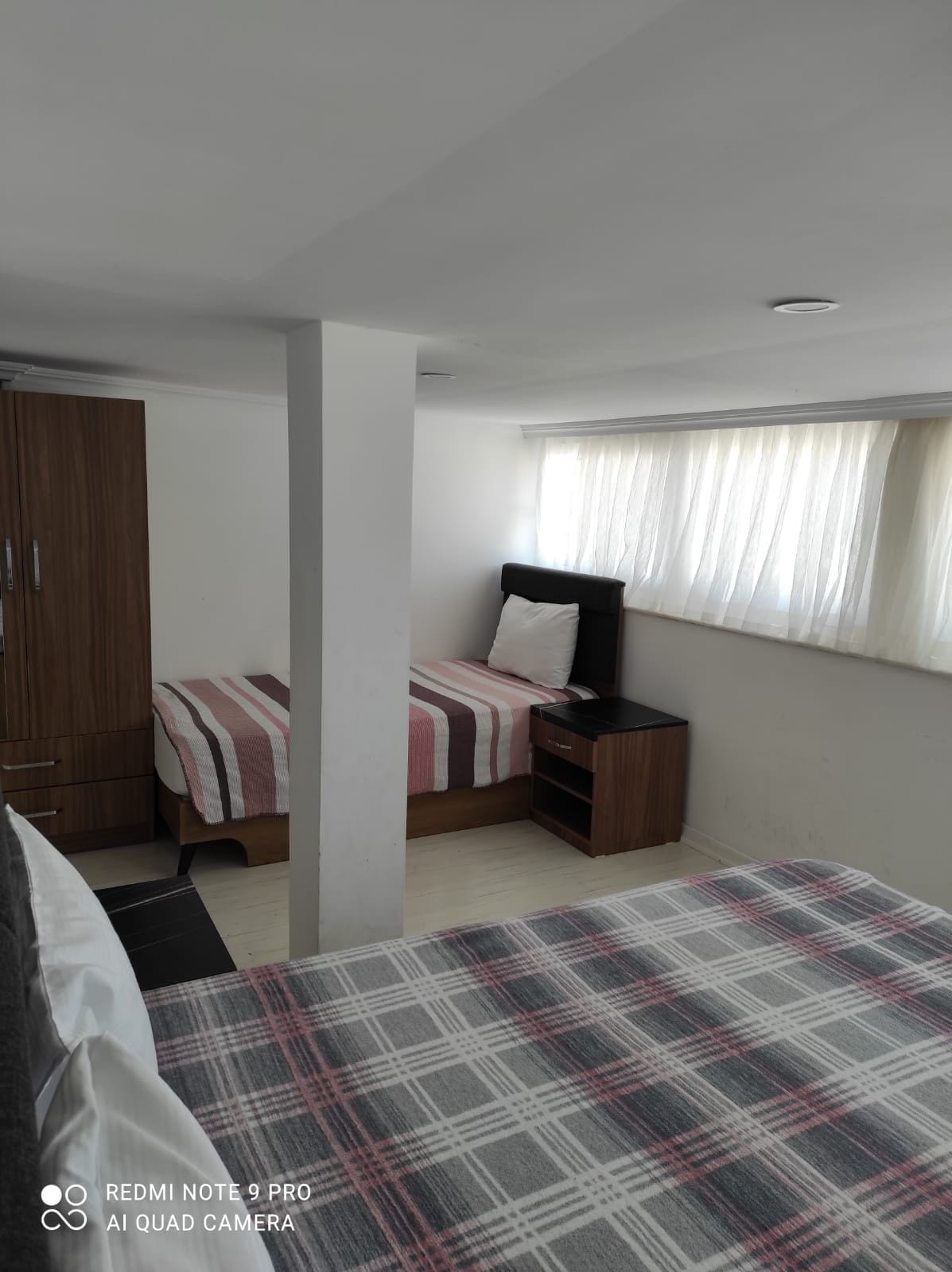 A well-lit bedroom area features two beds, one with a striped coverlet and the other with a plaid blanket. A bedside table is positioned next to the first bed, with natural light filtering in through the sheer window coverings. A wardrobe is also visible.