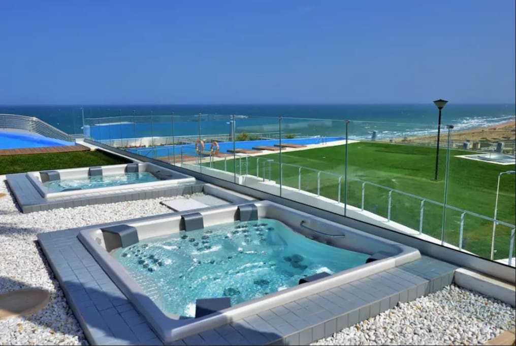 Some of the jacuzzis and swimming pools.
Piscinas climatizadas para adultos “Infinity pool”: Con una superficie de más de 200 m2 terminada en gresite e iluminación interior con cambios de color, dispone de escaleras para personas con movilidad reducida.
Dos jacuzzis climatizados con iluminación interior, con capacidad para 6-8 personas cada uno, realizados en material acrílico termoconformado de una sola pieza, con agarraderas en acero inoxidable, asientos ergonómicos y con impulsores tanto de agua como de aire, de la marca USSPA.
Duchas, tumbonas y sombrillas.
Plataformas con futones (colchonetas) en el ámbito de la piscina.
Piscina sur para adultos “Infinity pool”: Con una superficie de unos 230 m2 terminada en gresite e iluminación interior con cambios de color, dispone de escaleras para personas con movilidad reducida.
Plataformas con tumbonas en el ámbito de la piscina.
Jacuzzi climatizado con iguales características a las de la piscina climatizada.
Piscina –mirador en forma de proa de barco que se proyecta sobre los bloques de primera línea disfrutando de vistas panorámicas al mar.
Lago infantil tematizado con una superficie aproximada de 160 m2 con juegos infantiles integrados en el agua con pavimento imitación playa.
Chill out con más de 100 m2 compuesto por 2 barras, una húmeda que se integra en la piscina y otra barra normal con mesas, sofás y sillones bajo la sombra de modernas velas y refrescantes jardineras. Grandes playas de piscinas con césped natural o artificial dependiendo de la zona, con acceso directo desde los pasos peatonales, comunicadas con los ascensores y escaleras de los bloques.