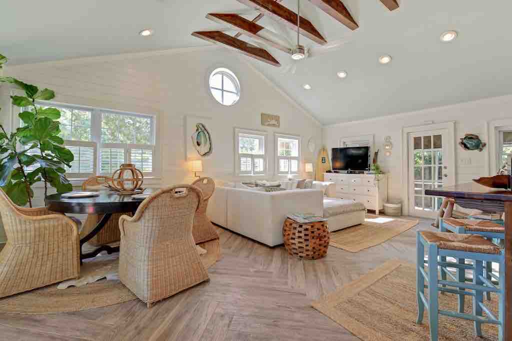 A light and airy living space features high vaulted ceilings adorned with wooden beams. Natural light fills the room through multiple windows. A round dining table is surrounded by woven chairs, while a comfortable sectional couch is positioned across from a sleek entertainment unit.