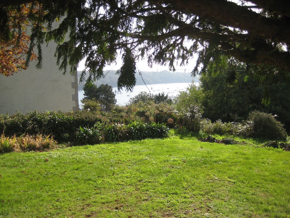 A green lawn is framed by overhanging branches, providing a view of the tranquil water. Various plants and shrubs are visible in the garden, while the distant shoreline hints at a serene landscape beyond.