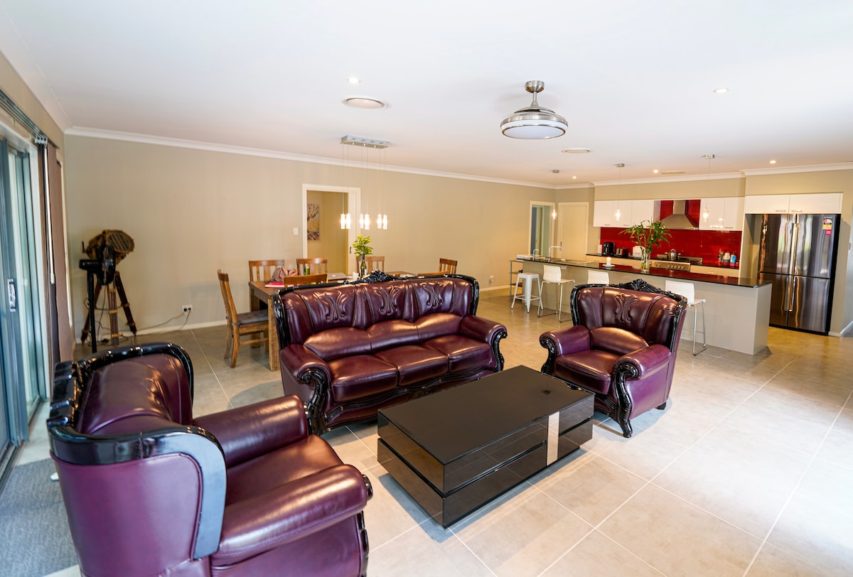 A spacious living area features deep purple leather sofas arranged around a dark coffee table. A dining table with chairs is positioned nearby, with a modern kitchen visible in the background, showcasing sleek cabinetry and stainless steel appliances.
