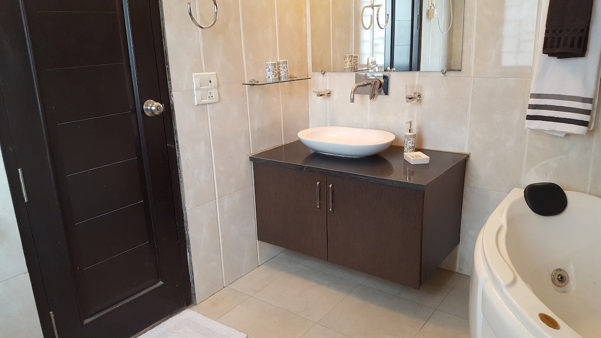 A modern bathroom is equipped with a sleek, wall-mounted sink set against light-colored tiles. A large mirror reflects the space, while a dark wood storage cabinet adds contrast. A spacious bathtub is positioned in the corner, and towel racks are conveniently placed nearby.