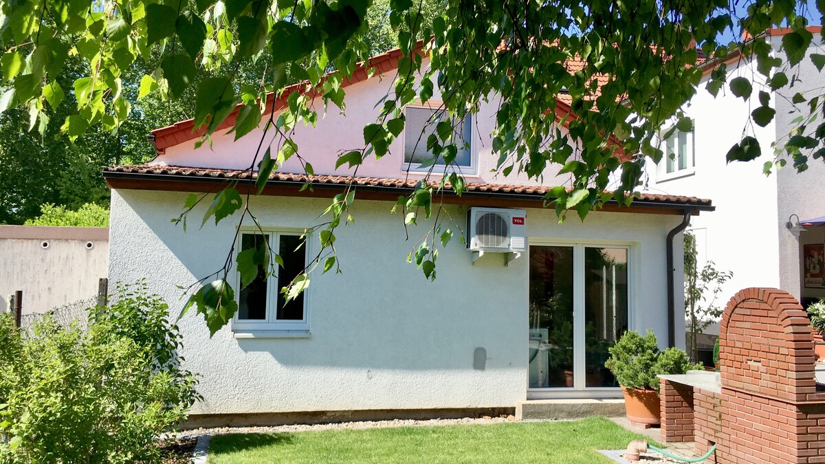 The exterior of the apartment is visible, featuring a light-colored façade and a terracotta roof. An air conditioning unit is mounted on the wall, and large sliding doors provide access to the interior. Green grass and small plants create a neat outdoor space.