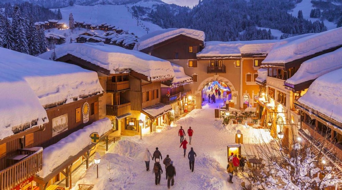 A snow-covered village square is illuminated by warm lights, with quaint buildings lining the path. Visitors stroll through the snowy landscape, enjoying the festive atmosphere. Fir trees dusted with snow stand nearby, enhancing the winter ambiance in this alpine setting.