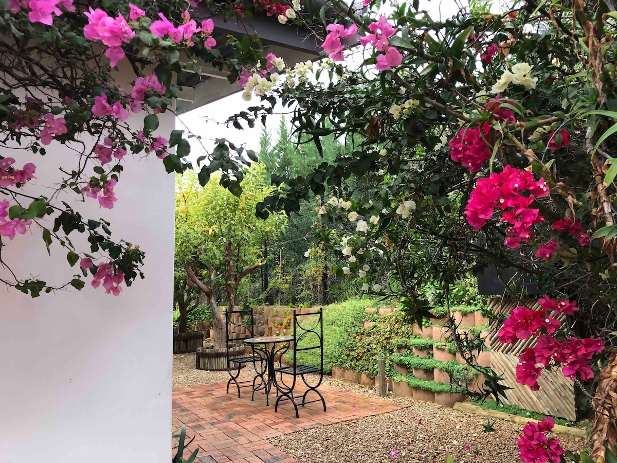 A private garden area features a small seating arrangement with a black table and wrought iron chairs. Colorful bougainvillea flowers frame the view, while a gravel pathway leads through lush greenery and landscaped plants.