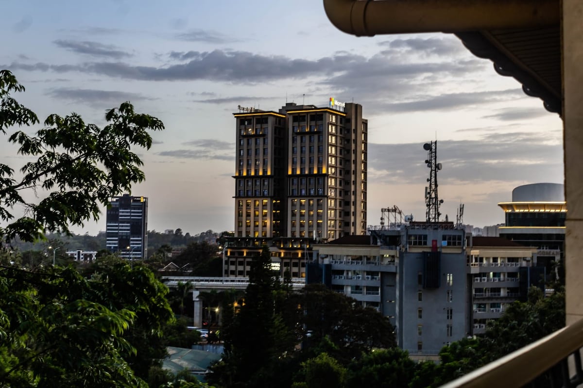 The image captures a view of urban Nairobi at dusk, showcasing a cluster of modern buildings. Towering structures with soft lighting contrast against a colorful sky, while greenery from nearby trees frames the scene, highlighting the apartment's connection to the city and nature.