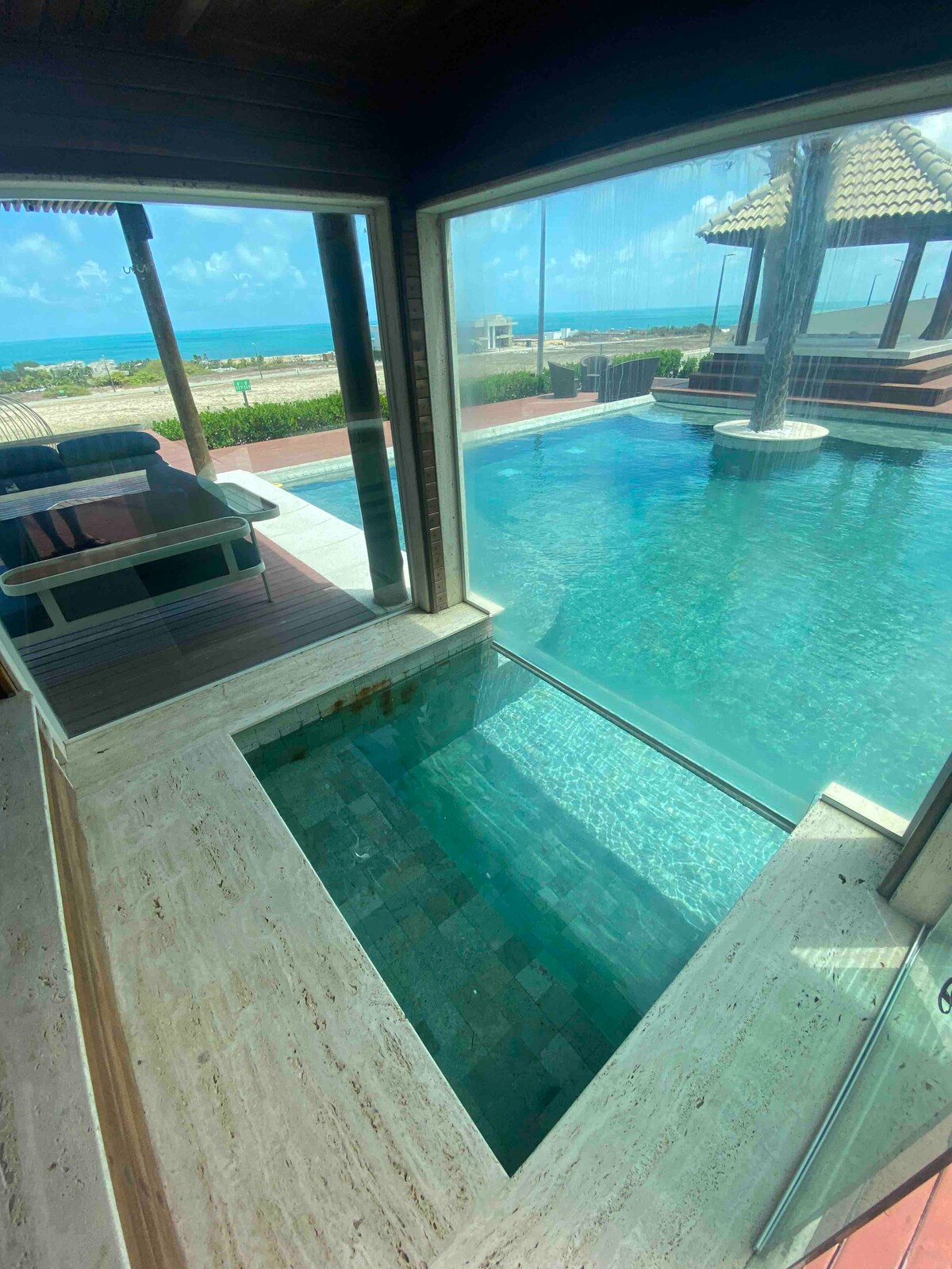 A serene outdoor area features a clear swimming pool adjacent to a wooden deck. A jacuzzi is visible by the poolside, and a shaded seating area adds comfort. The ocean can be seen in the distance, along with bright blue skies and scattered clouds.