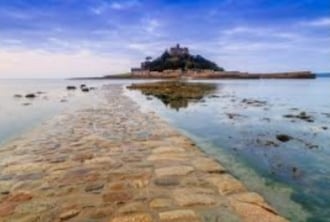 A wide view captures the rugged coastline leading to a small island topped with a historic structure. Calm waters reflect the sky, with smooth stones forming a path across the tidal flats. Soft clouds and varying shades of blue fill the sky above.