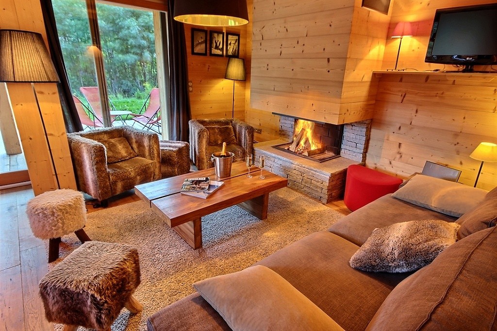 A cozy living room features a warm fireplace surrounded by wooden panels. Two plush armchairs and a spacious sofa with cushions offer comfortable seating. A wooden coffee table sits in the center, complemented by stylish side tables. Large windows provide a view of the outdoor space.