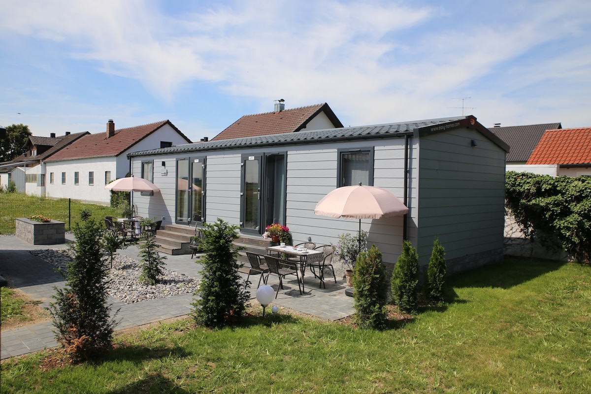 A well-maintained mobile home features large glass doors and an outdoor terrace. Two tables with umbrellas are positioned on the patio, surrounded by neatly arranged shrubs and greenery. The tranquil setting is complemented by a clear blue sky.