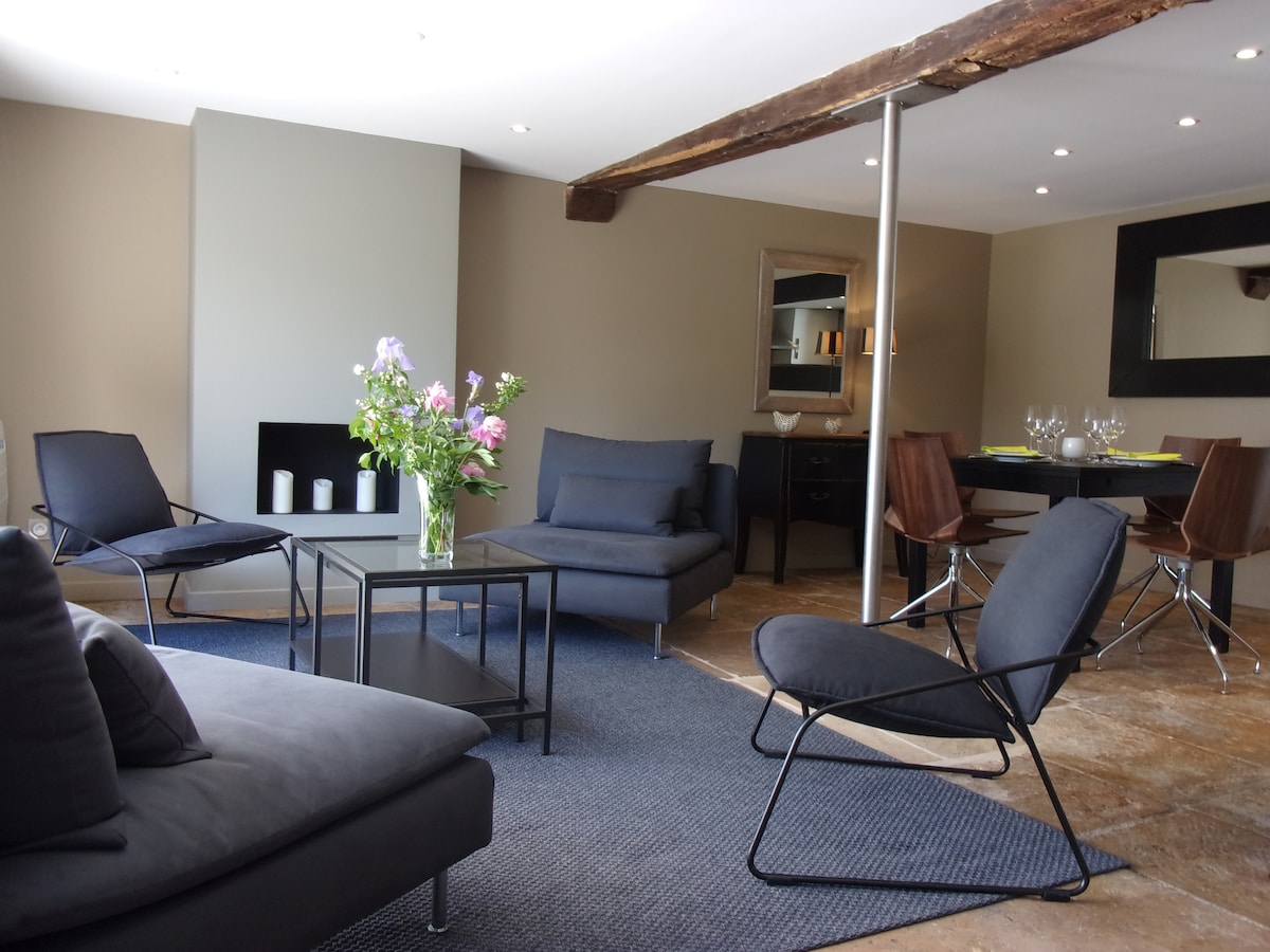 A spacious living area is furnished with modern seating arrangements, including two lounge chairs and a comfortable sofa. A sleek coffee table sits in the center, complemented by a floral arrangement. The open layout transitions into a dining area featuring a dark table set for dining.