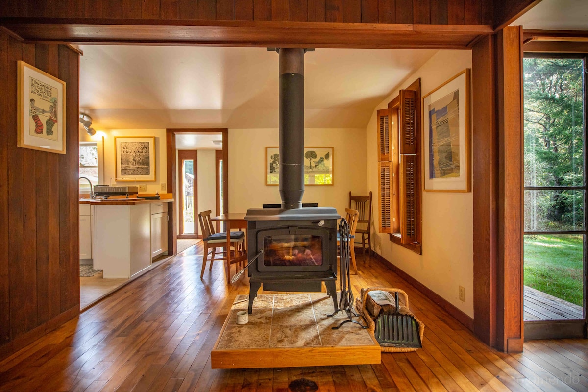 An inviting living space features a central wood stove on a stone platform, surrounded by warm wood paneling. Natural light enters through large windows and glass doors, illuminating rustic furnishings, including chairs and a dining table, with glimpses of the lush garden beyond.