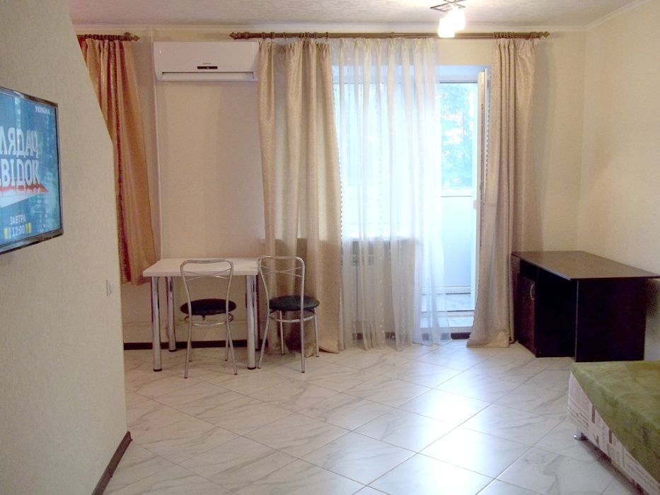 A light-filled living space is furnished with a simple table and chairs positioned near a window, draped with sheer curtains. A television is mounted on the wall. A dark cabinet is visible beside a bed with an unmade cover, set against a neutral-toned wall.