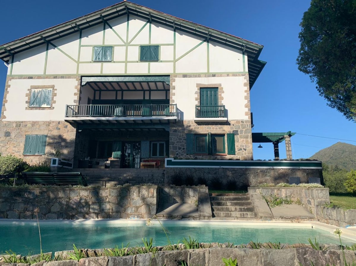 The spacious two-story house features a stone facade with green shutters. A covered balcony extends across the front, providing shade. Outside, a long pool is surrounded by greenery, creating a calm outdoor space. The backdrop showcases tranquil hills under a clear sky.