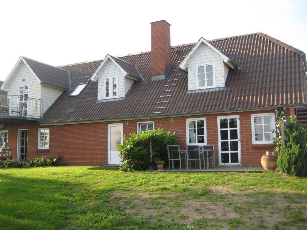 A two-story house features a mix of brick and wood siding, accented by a sloped roof. A patio area is visible with a table and chairs, surrounded by well-maintained greenery. Windows provide natural light, and a small balcony is seen on the upper level.