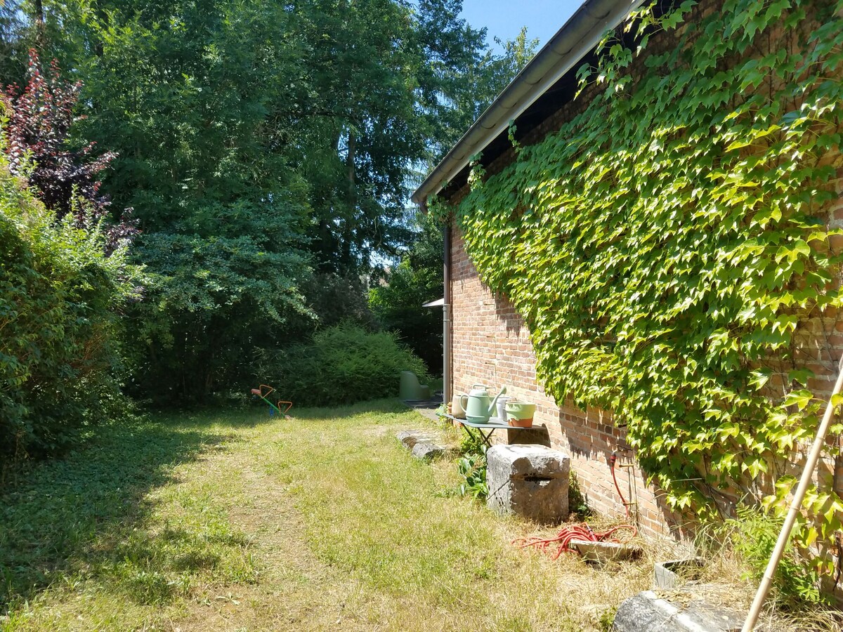 A lush garden space is featured, bordered by greenery and plants. A brick wall is partially covered with climbing vines. Several pots are placed along a stone surface, and a pathway leads through the grassy area, with trees providing a natural backdrop.