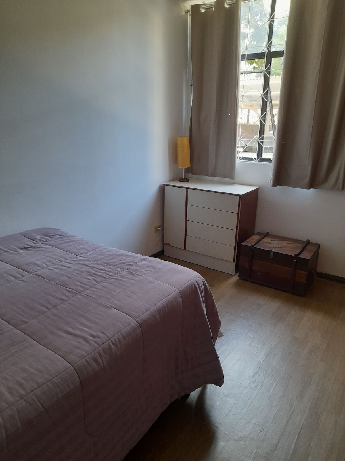 The comfortable bedroom showcases a spacious bed draped in a soft purple coverlet. A wooden dresser with multiple drawers stands beside the bed, while a small trunk adds character. Natural light enters through a window framed by soft curtains.