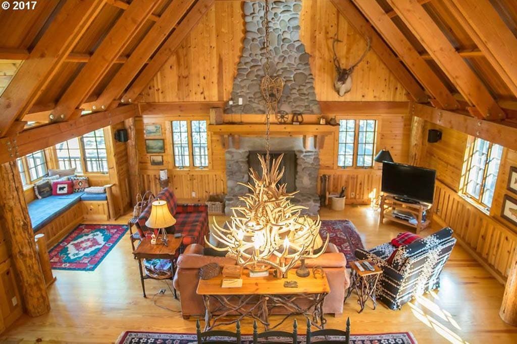 The great room features soaring vaulted ceilings and rich wood walls, complemented by large windows that invite natural light and showcase scenic views. A prominent river rock fireplace dominates the space, while a large antler chandelier and rustic furnishings add to the lodge-like charm.