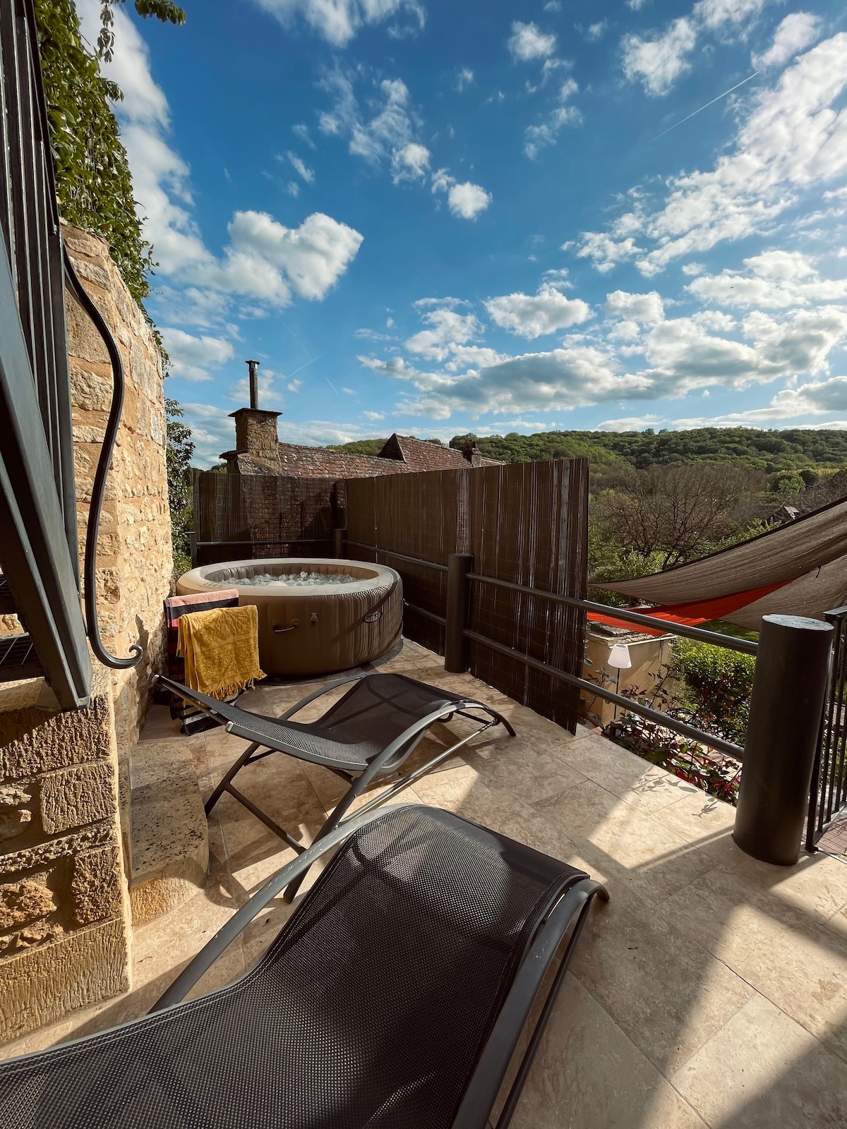 An outdoor terrace features a bubbling hot tub surrounded by scenic views of the countryside. Two lounge chairs provide a comfortable space for relaxation, while a textured blanket is draped over one chair. A hammock adds to the inviting ambiance of the area.