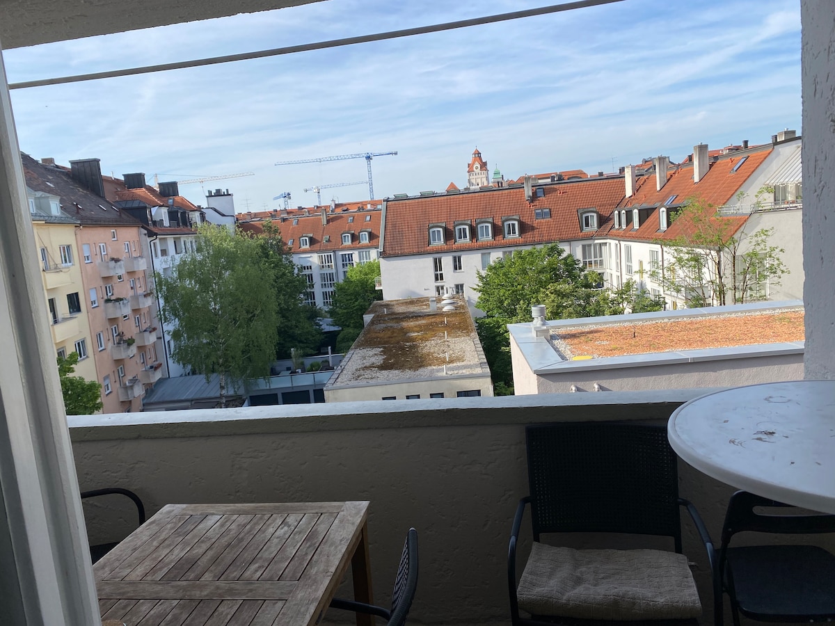 A balcony view captures the surrounding neighborhood, featuring rooftops with red tiles and greenery visible below. A wooden table and two black chairs are arranged on the balcony, providing a space for outdoor relaxation. The sky is bright with scattered clouds.