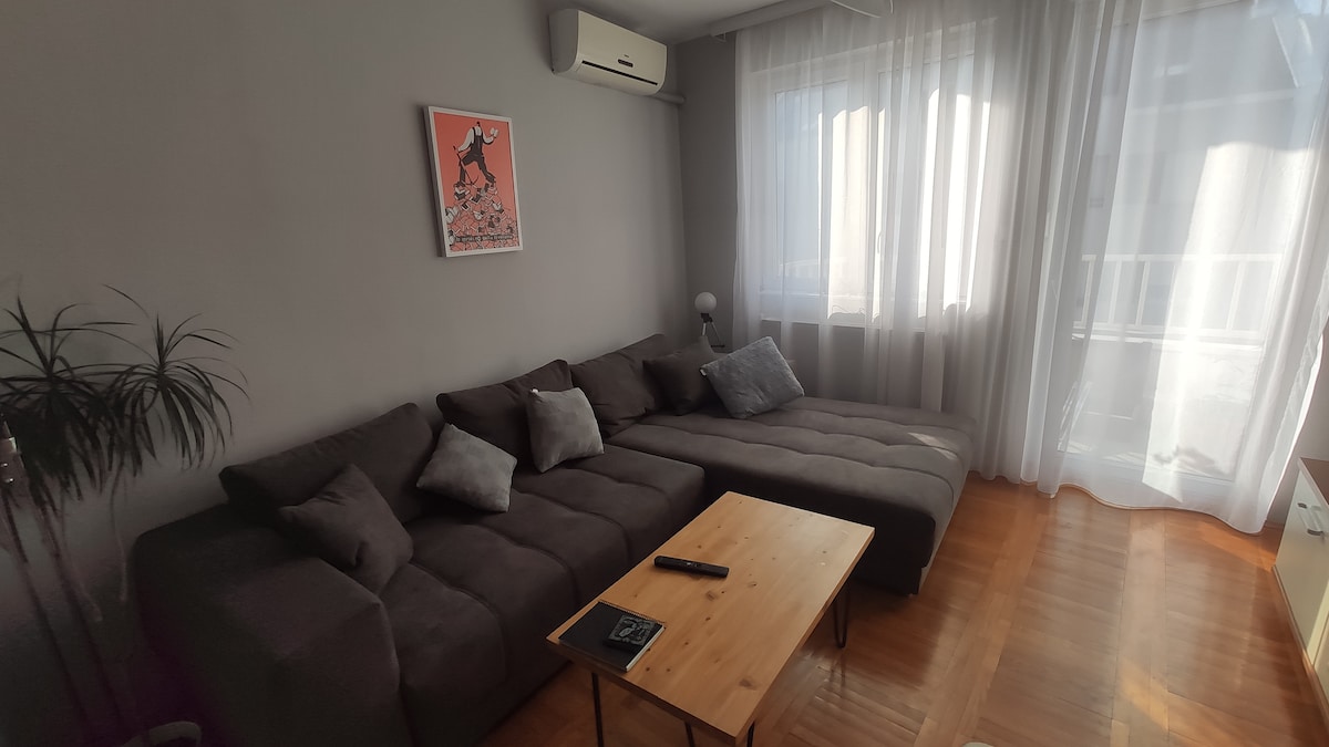 A spacious living room is furnished with a comfortable sectional couch, accented by several throw pillows. A light wood coffee table is positioned in front, with a remote control resting on it. Large windows draped in sheer curtains allow natural light to fill the room.