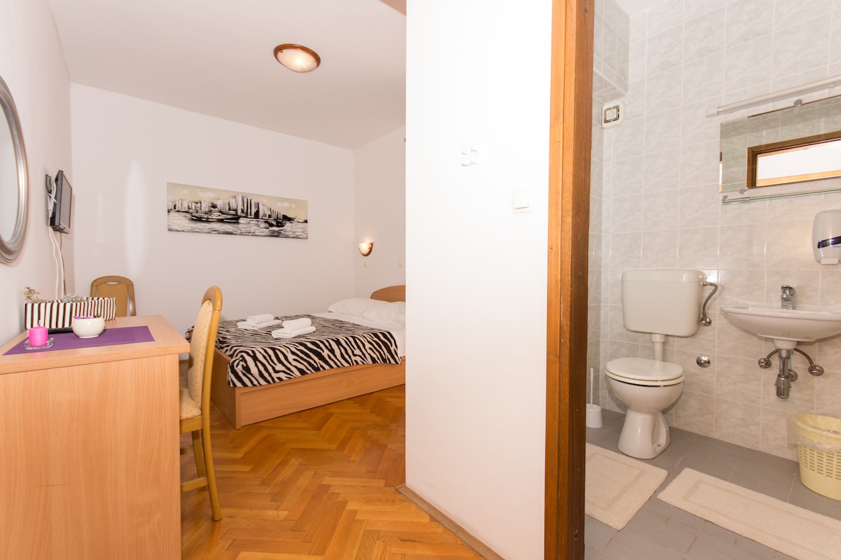 A spacious room is presented with a bed featuring a zebra-striped coverlet and a simple wooden chair. A small desk with a mirror is visible. An adjoining bathroom with basic amenities is accessible, showcasing white tiles and wall fixtures.