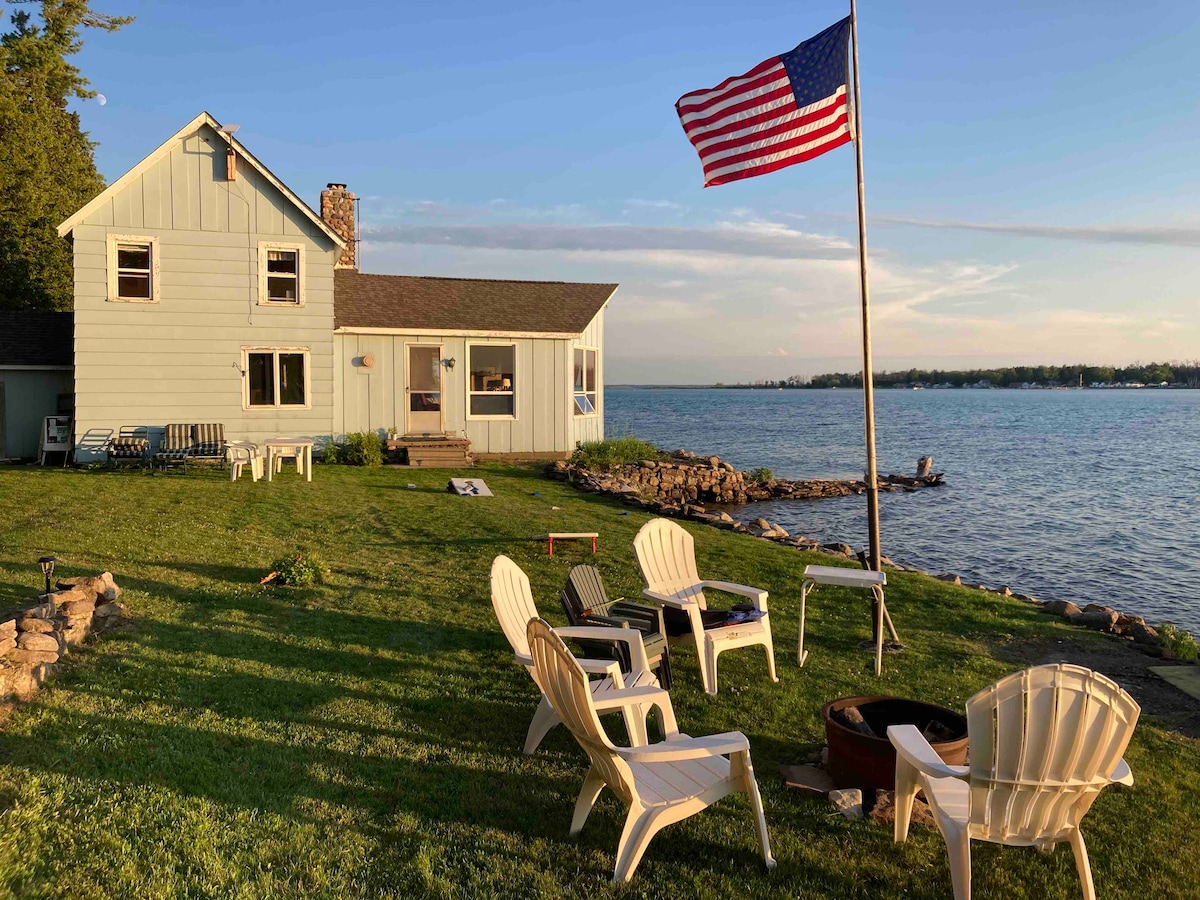 A light blue cabin is positioned along the water's edge, complemented by a grassy area featuring several white Adirondack chairs. A flagpole proudly displays the American flag. The serene river view enhances the peaceful ambiance of the setting.