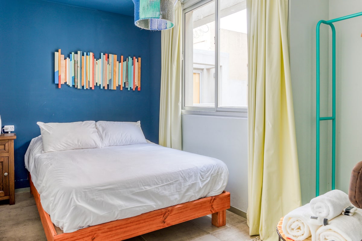 A bright sleeping area features a spacious bed with white linens against a rich blue wall. A colorful wall art piece made of wooden planks adds character. Natural light flows through a large window, accompanied by soft yellow curtains that frame the view.