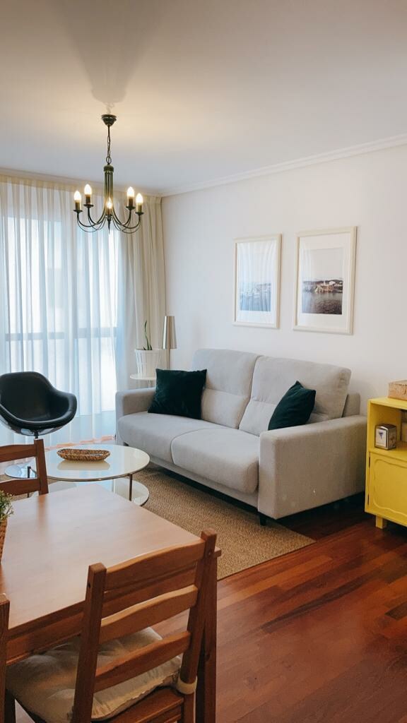 A living area features a comfortable sofa with dark cushions, a round glass coffee table, and a yellow side cabinet. Two framed artworks adorn the wall, and sheer curtains allow natural light to filter in, creating a peaceful ambiance in the space.
