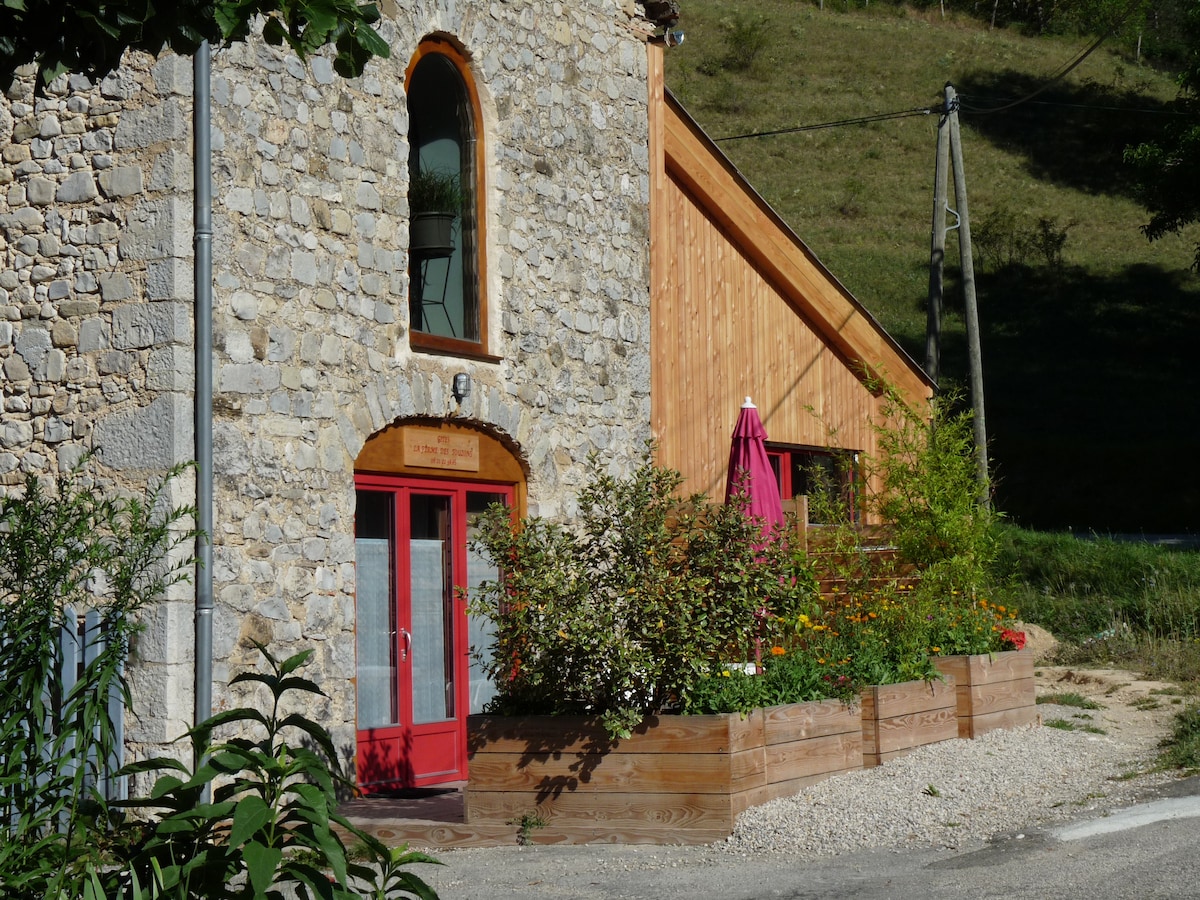 The exterior view features a charming blend of stone and wood architecture. A vibrant red door stands as the entry point, accompanied by a flowering garden and a shaded seating area with a pink umbrella. The natural landscape provides a serene backdrop.