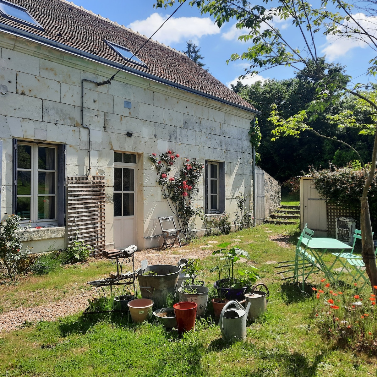 The exterior of the stone cottage is visible, featuring two large windows with flowerbeds below. A small dining table and chairs are placed on the lawn, surrounded by various potted plants. A pathway leads to the entrance, with greenery and trees in the background.