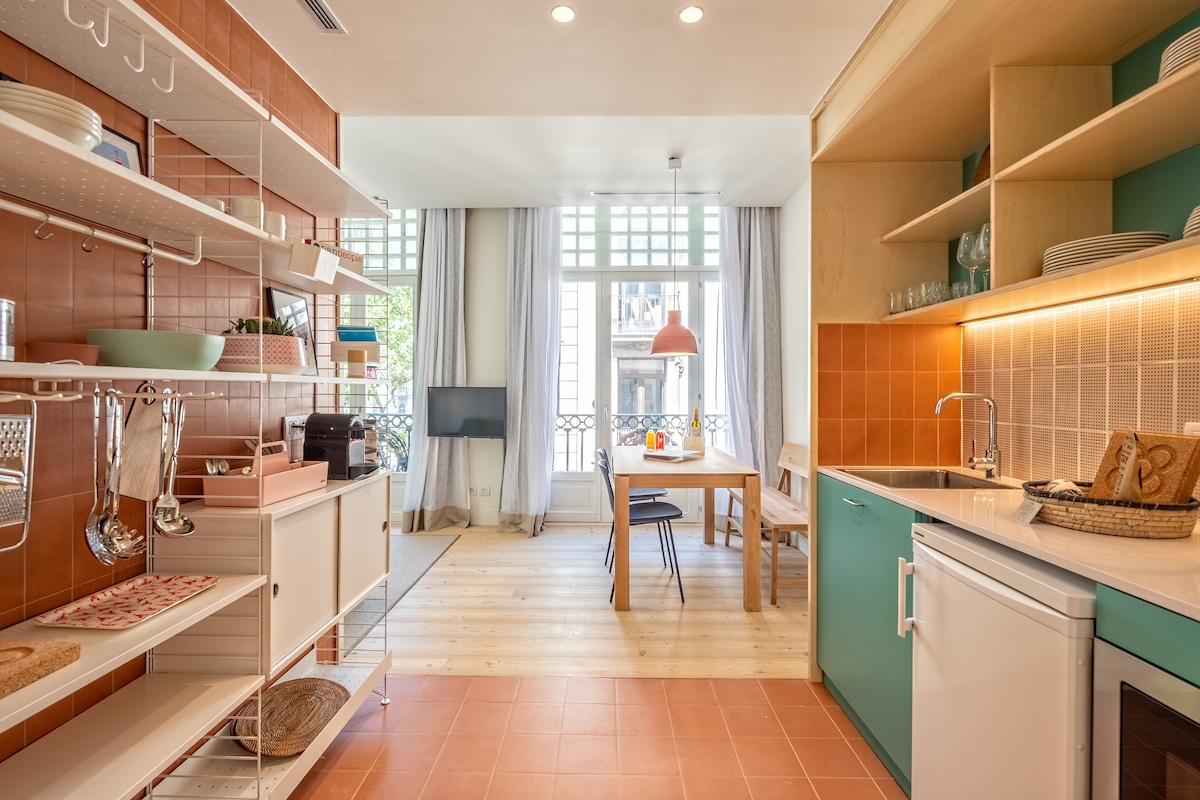 A well-equipped kitchen is shown with a mix of open shelving and cabinetry. Natural light floods the space through large windows, illuminating a dining area with a wooden table and chairs. The color palette features warm oranges and greens, creating a harmonious atmosphere.