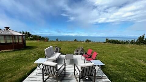 An outdoor seating area is arranged on a wooden deck, featuring several chairs and a central table. The expansive grassy area surrounds the setting, with views of the sea and distant mountains under a partly cloudy sky.