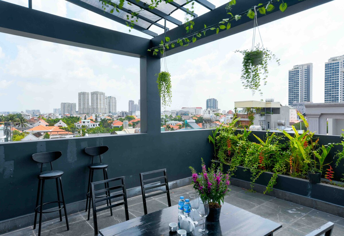 A spacious rooftop area is presented with seating options, including bar stools and a table. Green plants and flowers add natural elements, while the city skyline offers a panoramic backdrop. The space is designed for relaxation and enjoying scenic views.