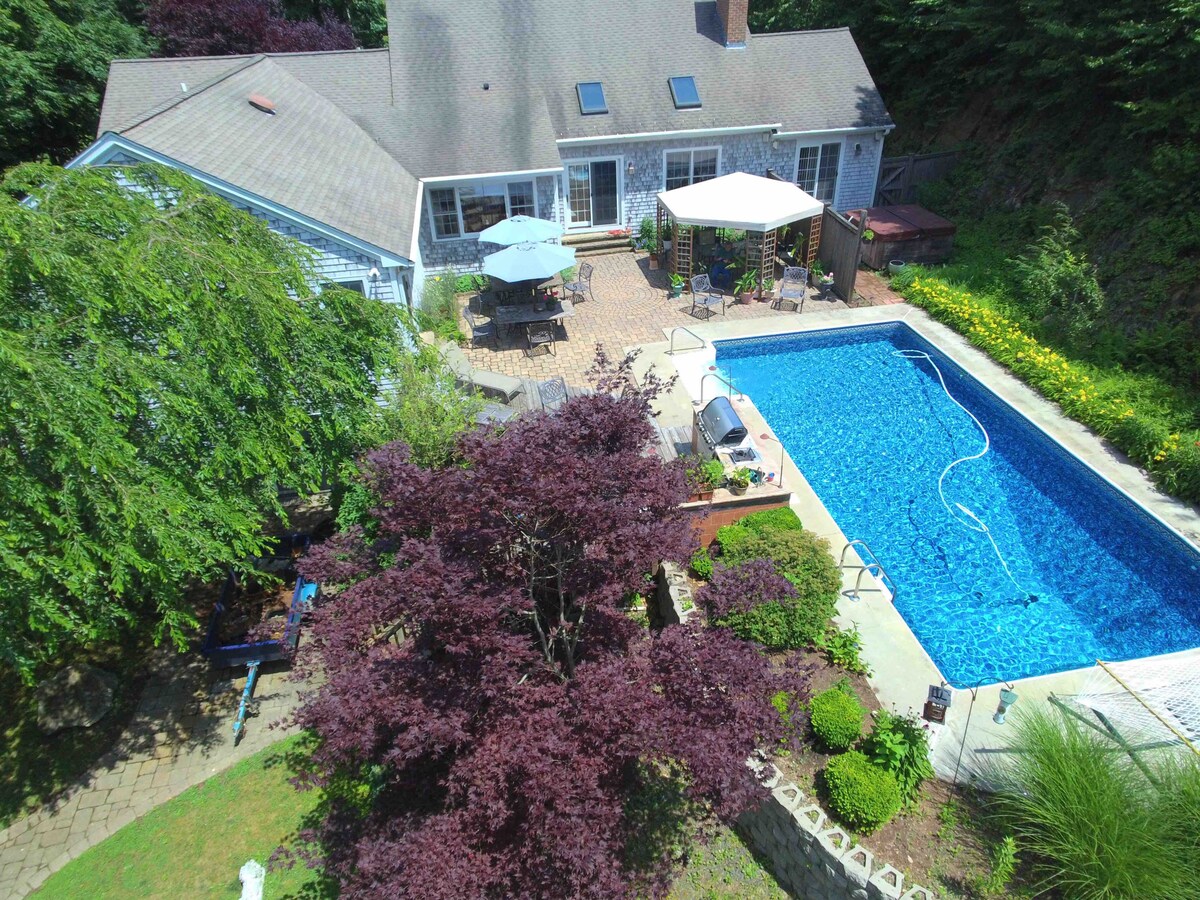 An aerial view captures a private pool surrounded by a stone patio and vibrant landscaping. Sun loungers and an umbrella-shaded table are visible near the water, while a grill is positioned nearby. The home is adorned with large windows, allowing light to fill the interior.