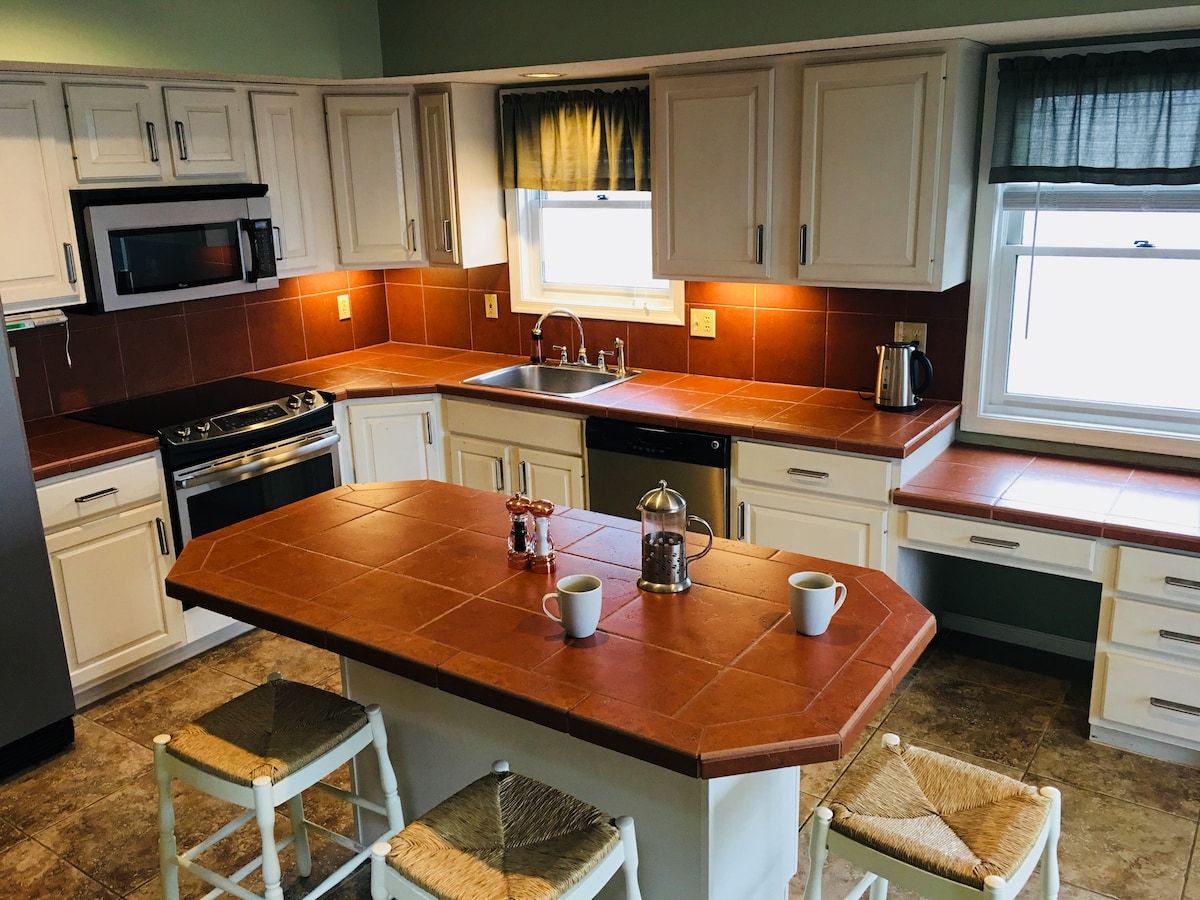 The kitchen features a central island with tiled countertop and seating for three. Modern appliances include a sleek stove and microwave. Natural light pours in through a window, complemented by a neutral color scheme and warm-toned cabinetry, creating a functional and welcoming cooking space.