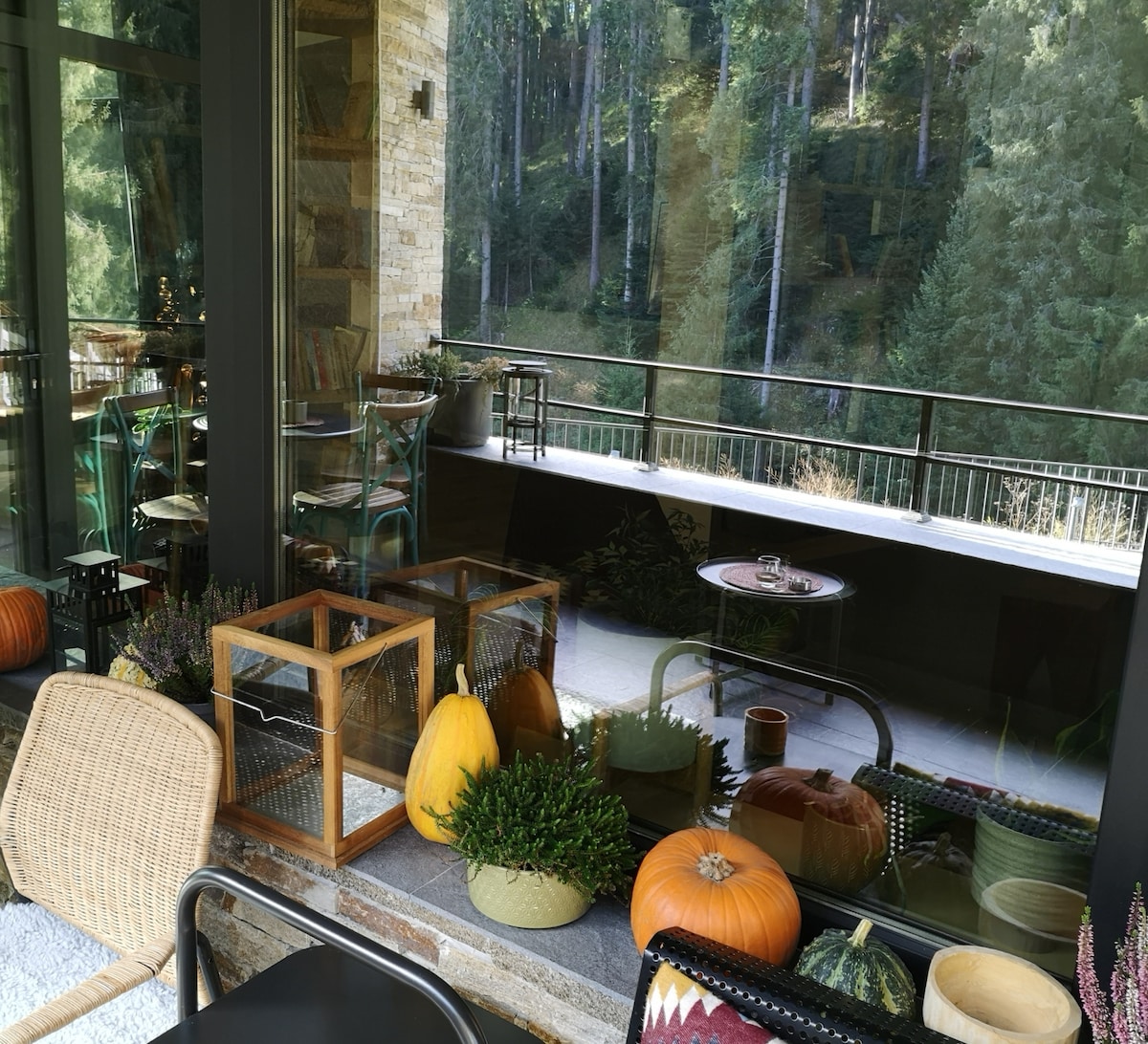 A spacious terrace is visible, featuring various pumpkins and decorative plants along the edge. The glass railing provides an unobstructed view of the surrounding forest. Tables and chairs are arranged for outdoor seating, contributing to a welcoming outdoor environment.