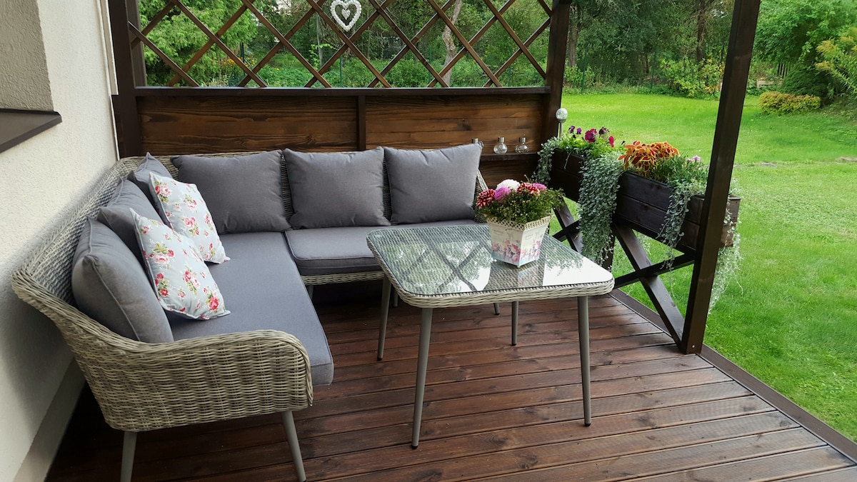 A shaded terrace features a comfortable seating area with a gray sofa adorned with floral pillows. A glass table sits in front, complemented by a potted plant. Lush greenery and blooming flowers can be seen in the background, creating a peaceful outdoor retreat.