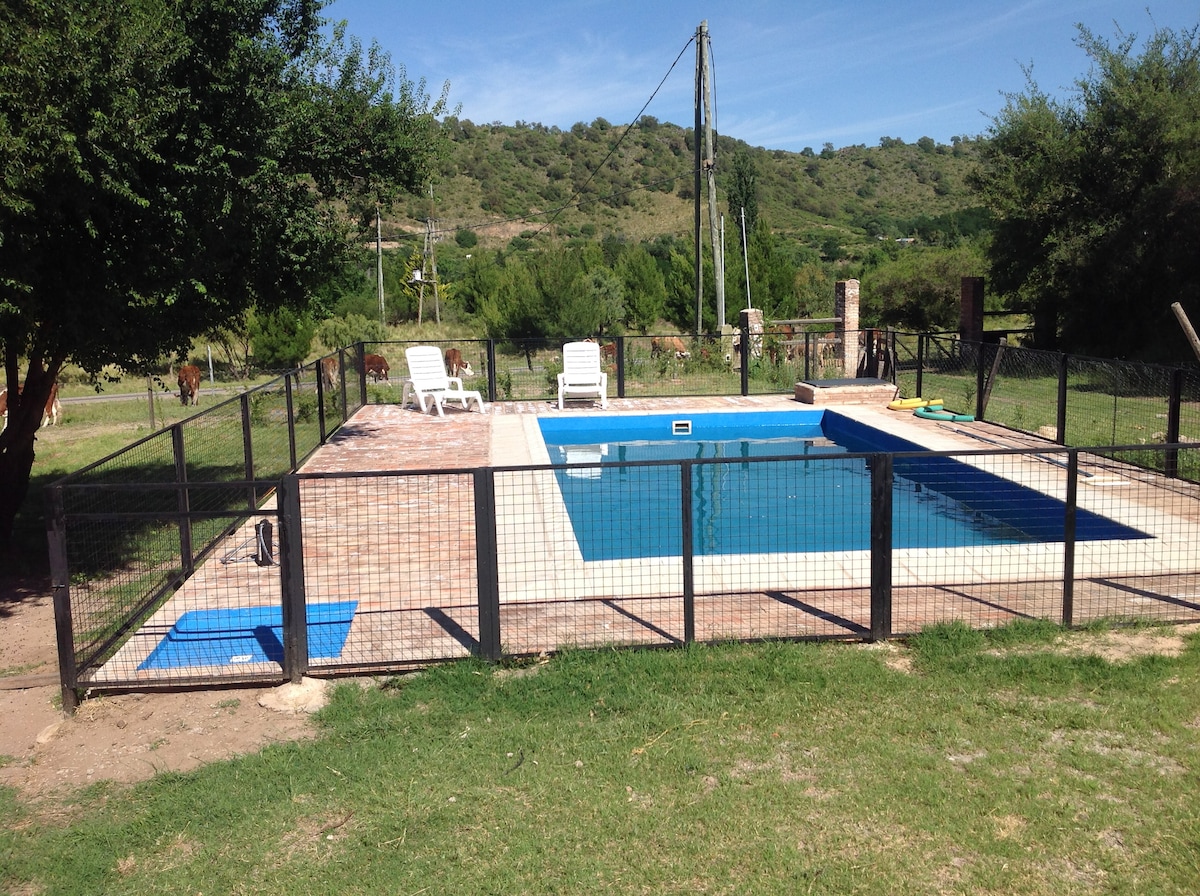 An outdoor swimming pool is surrounded by a sturdy fence, ensuring safety. Several lounge chairs are positioned on the pool deck. Lush greenery and hills are visible in the background, complementing the serene landscape. Bright blue water reflects the clear sky above.