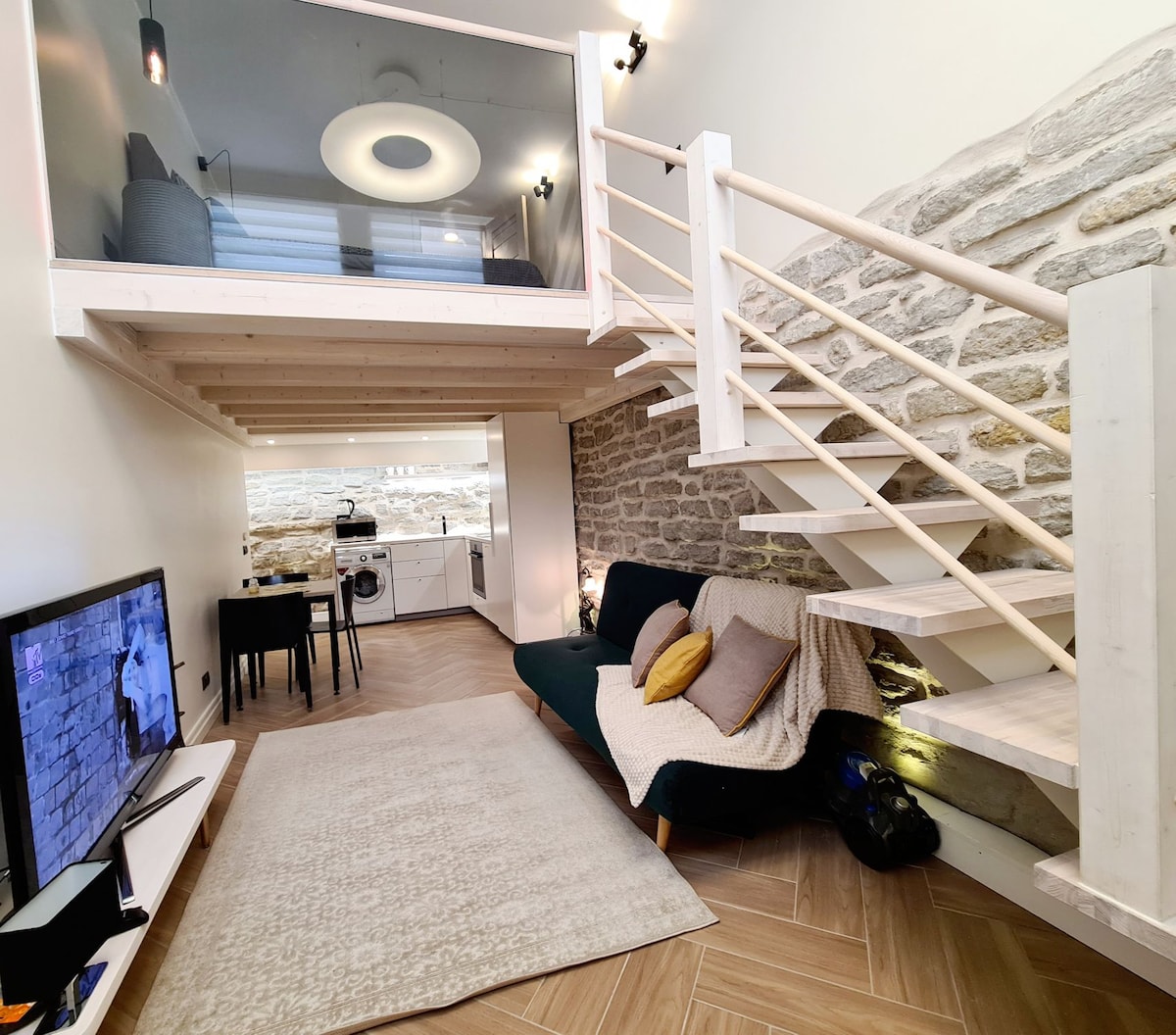 A spacious living area features a staircase leading to a loft, with exposed old stone walls and soft lighting. A green sofa is paired with a light rug, while a television and dining table are positioned nearby. The open kitchen is visible in the background.