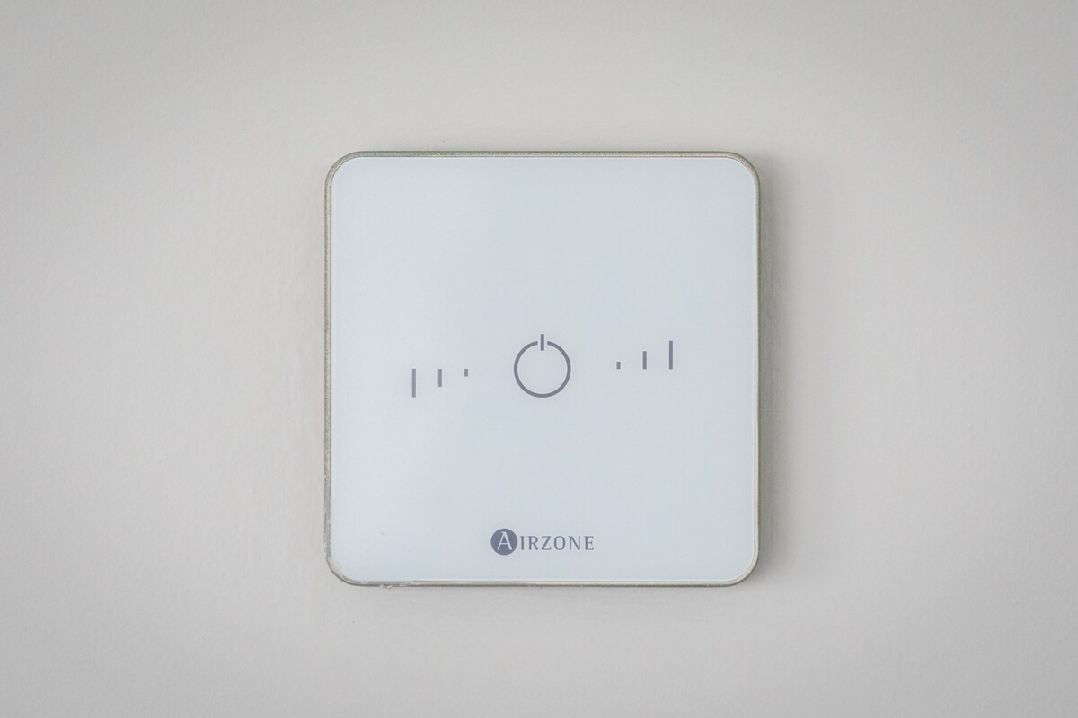 A thermostat is mounted on a light-colored wall, featuring a circular dial at the center with indicators around it. The minimalist design and modern branding suggest user-friendly temperature control.