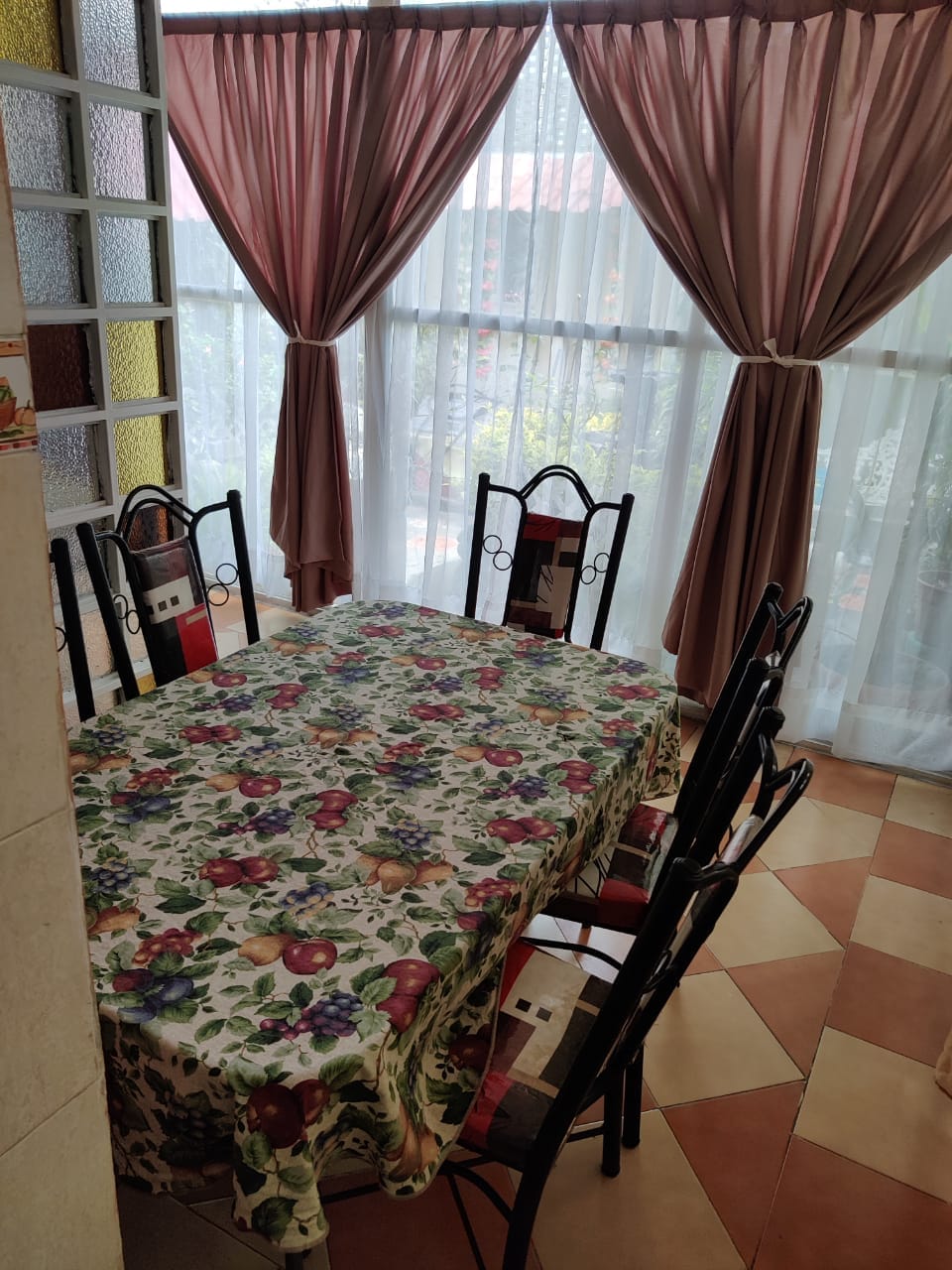 A dining area features a large table covered with a floral tablecloth, surrounded by six black chairs. Natural light filters through sheer white curtains, which are complemented by soft pink drapes. A view of greenery is visible through the windows.