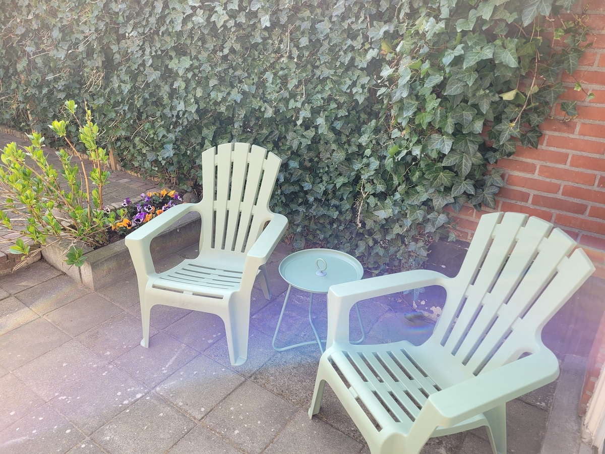 A small outdoor seating area features two light-colored lounge chairs and a minimalist round table. Lush green ivy covers the wall behind, with colorful flower beds adding vibrancy to the space. The stone patio provides a cozy atmosphere for relaxation.