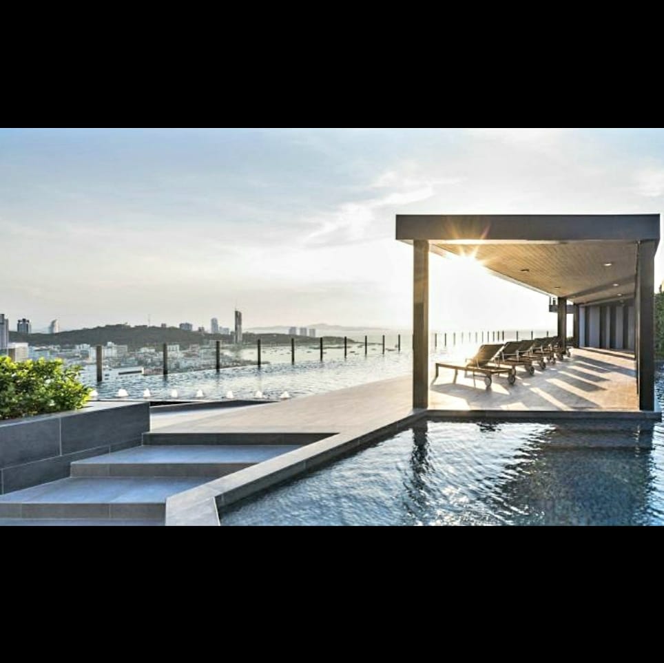 An infinity swimming pool is bordered by sleek lounge chairs, overlooking a panoramic view of the city skyline. Soft sunlight reflects off the water's surface, creating a serene atmosphere. The terrace is enclosed by glass railings, emphasizing the expansive scenery.