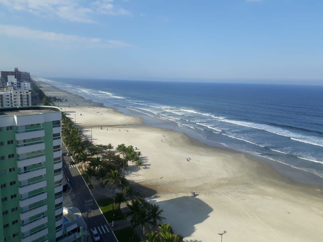 A panoramic view of a vast sandy beach meets the ocean, with gentle waves lapping at the shore. Palm trees line the beachfront, and a broad pathway runs alongside the beach, facilitating access. The image features a clear blue sky, enhancing the beach's serene ambiance.