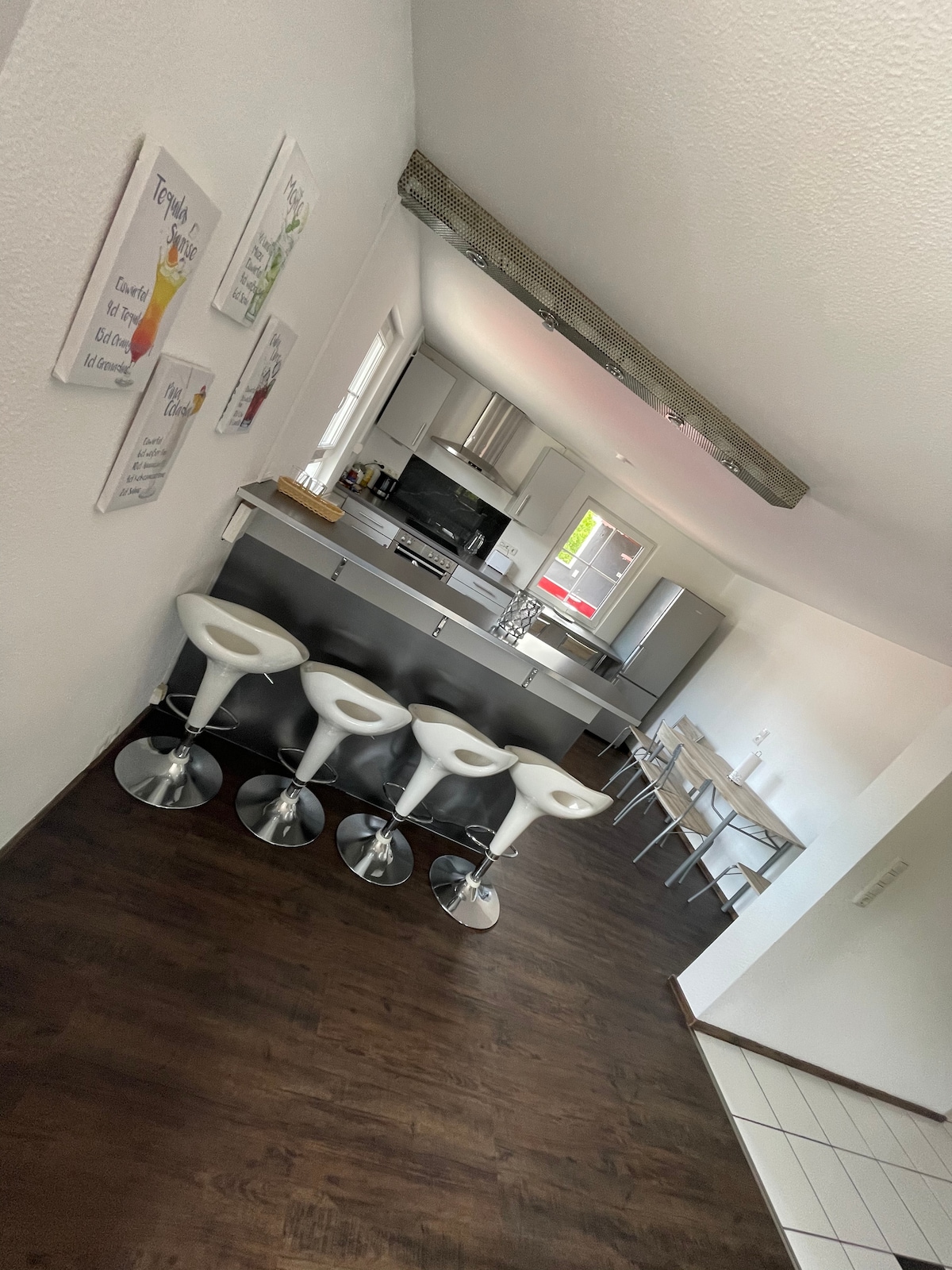 The kitchen area features modern appliances with sleek cabinetry and a spacious countertop. Four white bar stools are positioned at the counter, complemented by a small dining table with metallic chairs. Light wood accents are visible in the ceiling beams, enhancing the overall design.